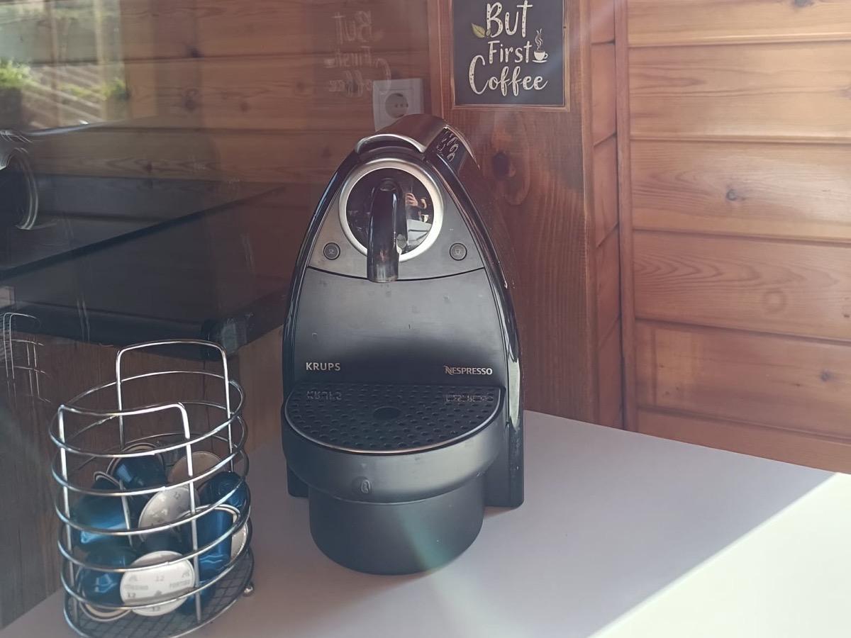 Cafetera de cápsulas sobre la encimera, con las cápsulas en un cestillo y un cartel de «But First Coffee» en la pared de madera