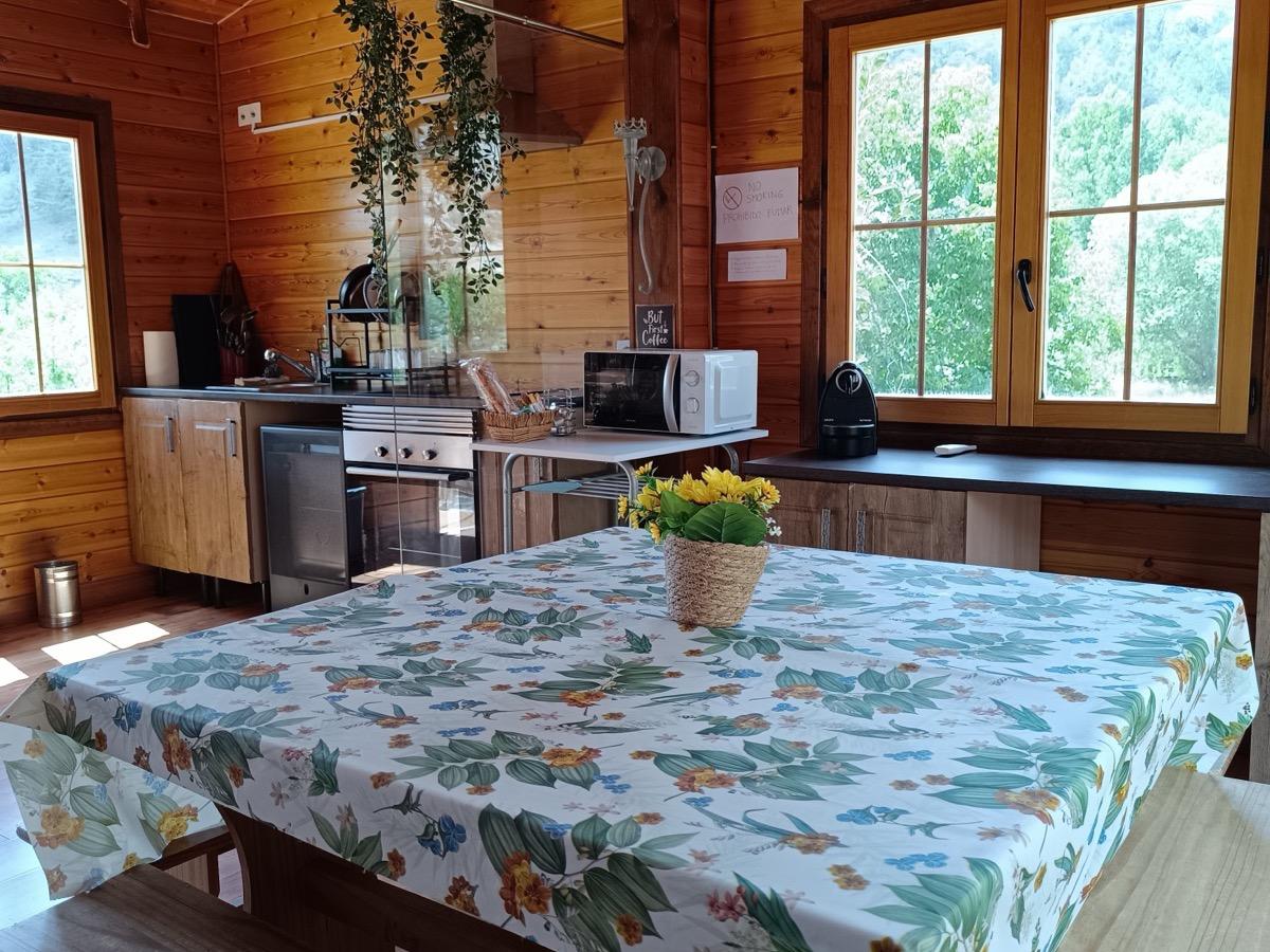 Mesa del comedor con mantel de flores y un ramo de flores amarillas, con la cocina y el techo abovedado de madera detrás