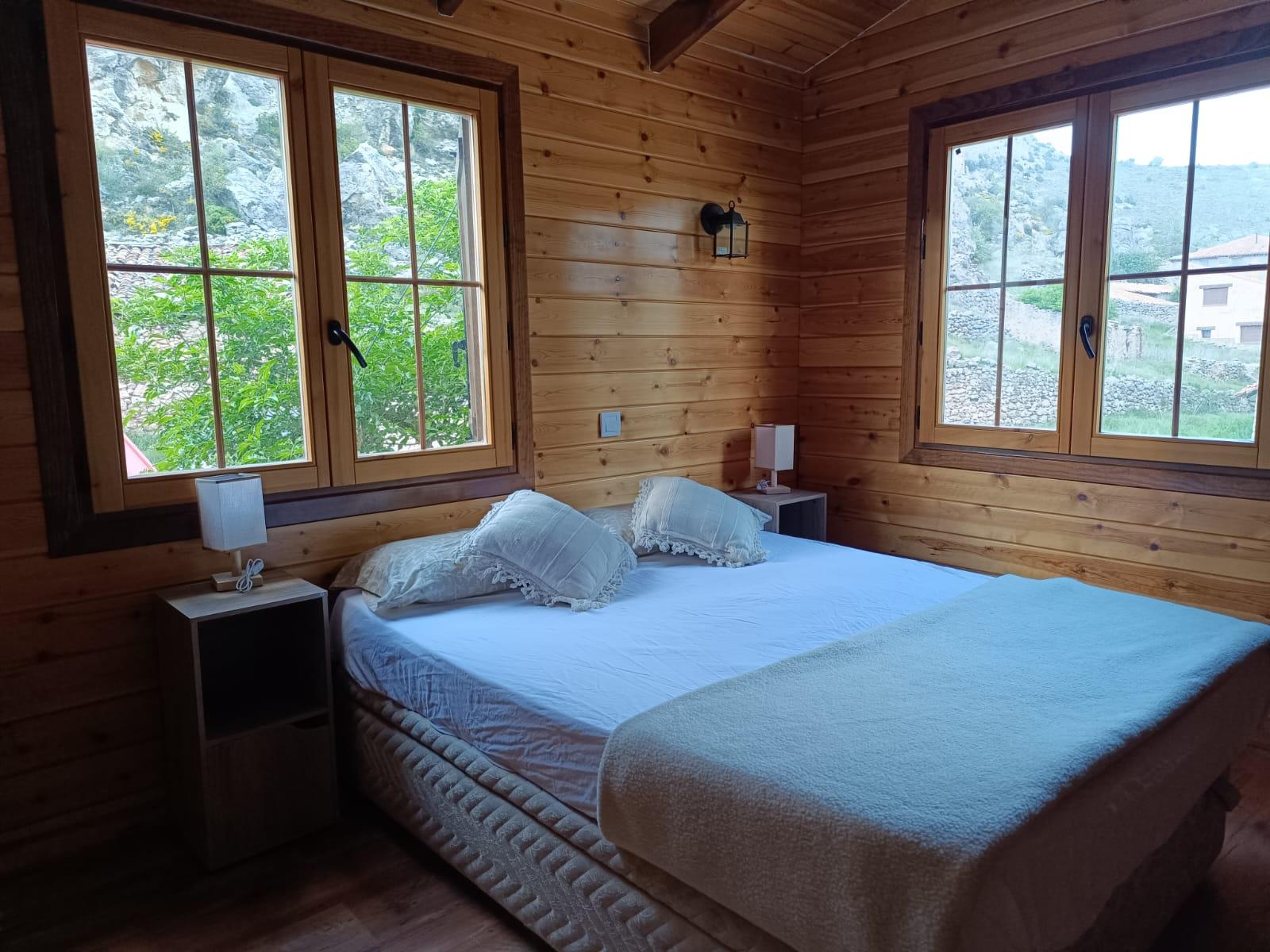 Habitación principal con cama de matrimonio entre dos ventanas grandes con vistas a la montaña y al pueblo
